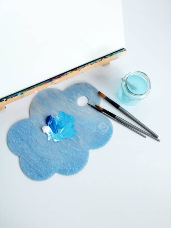 Flat lay shot of a canvas on an easel, mixed blue paint and paintbrushes with Cork & Chroma's Limited Edition Cloud Palette in Nimbus (blue sparkle)